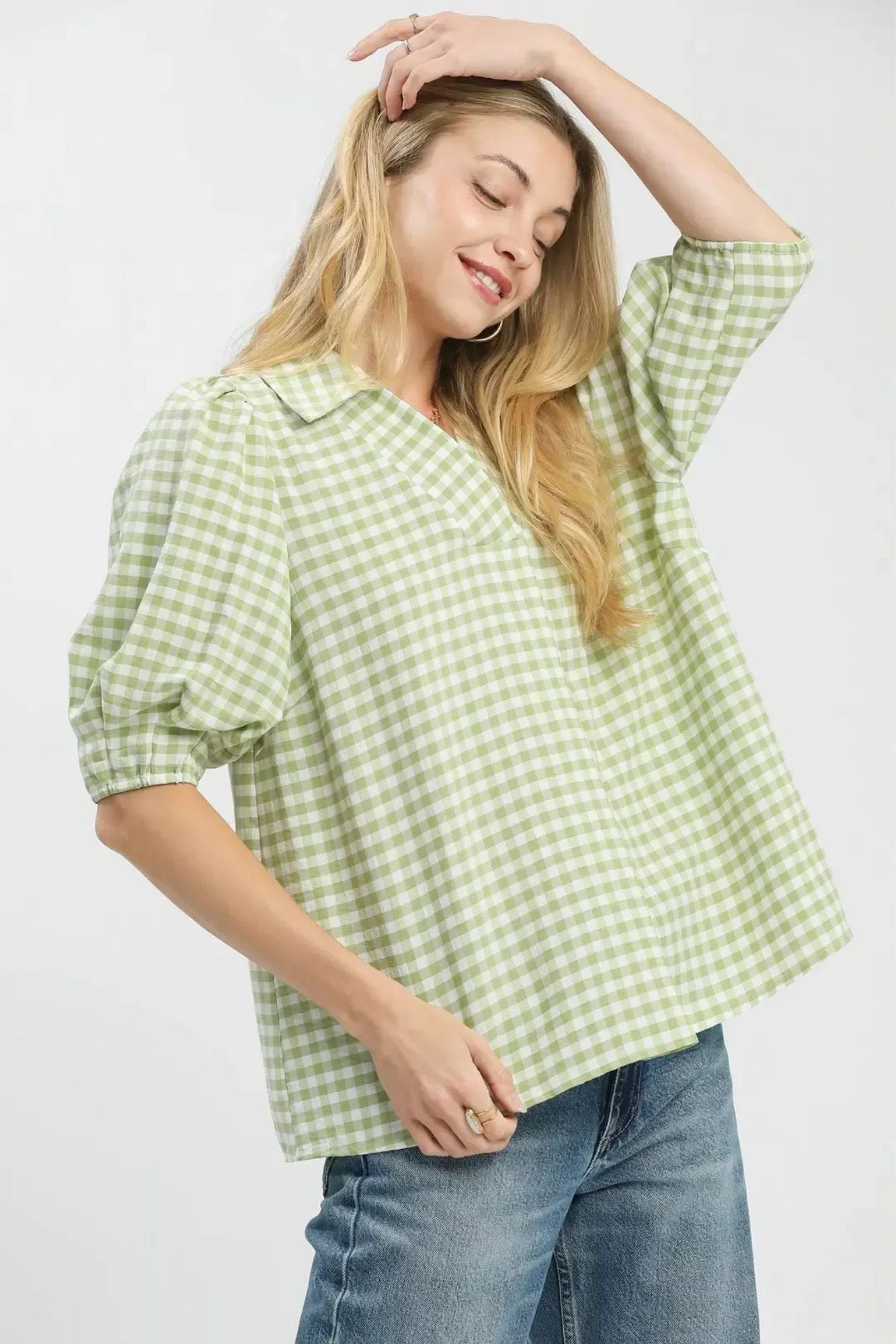 A woman wearing a light green and white gingham check short-sleeve blouse with puff sleeves and a collar, paired with blue jeans, against a plain white background.