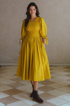 Woman wearing a yellow midi dress with smocked bodice, round neckline, and puffed sleeve standing in a room with tiled floor and beige walls.