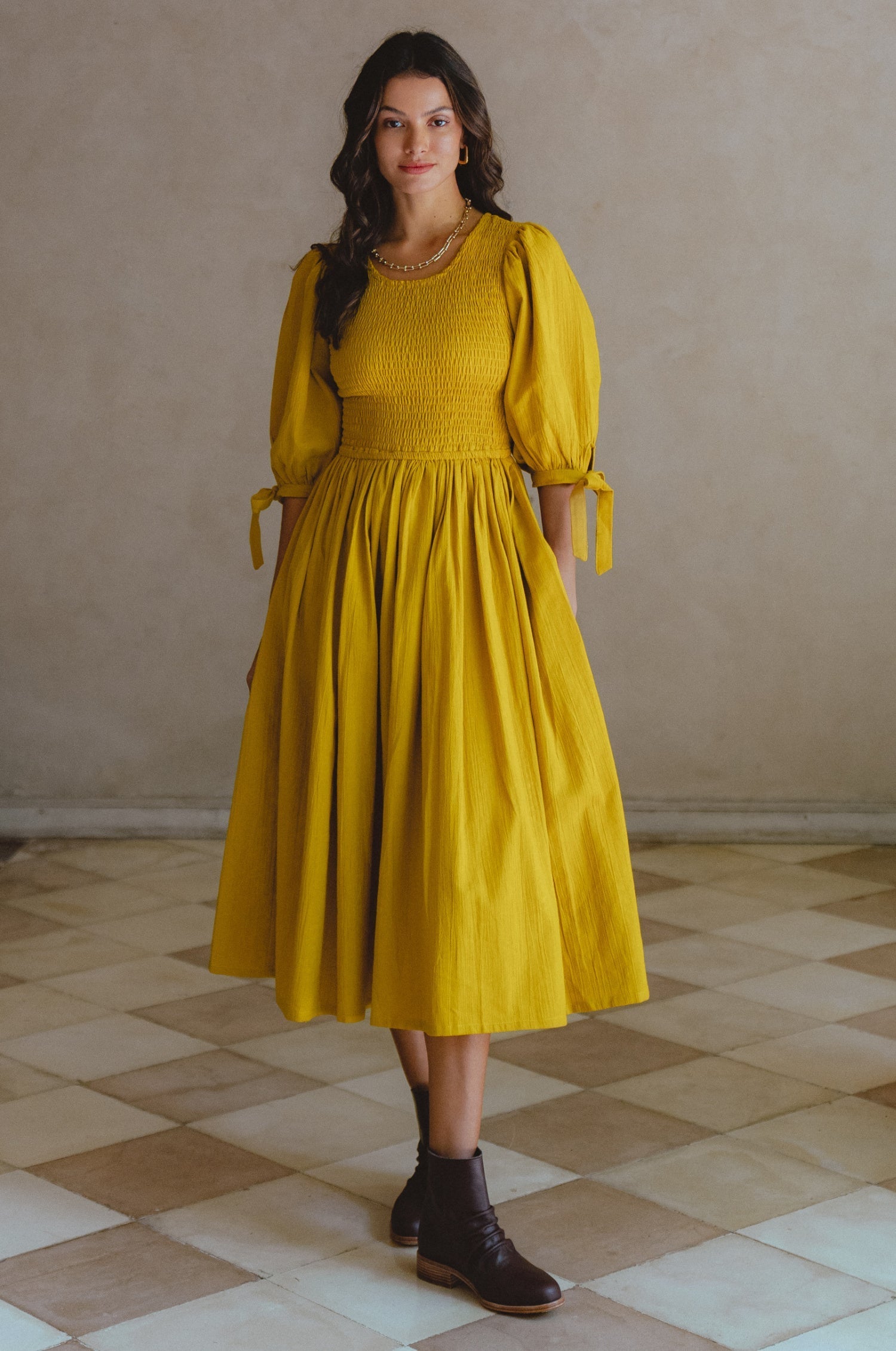 Woman wearing a yellow midi dress with smocked bodice, round neckline, and puffed sleeve standing in a room with tiled floor and beige walls.