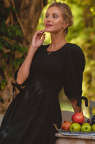 Woman in a black dress standing next to a table with fruits and vegetables outdoors.