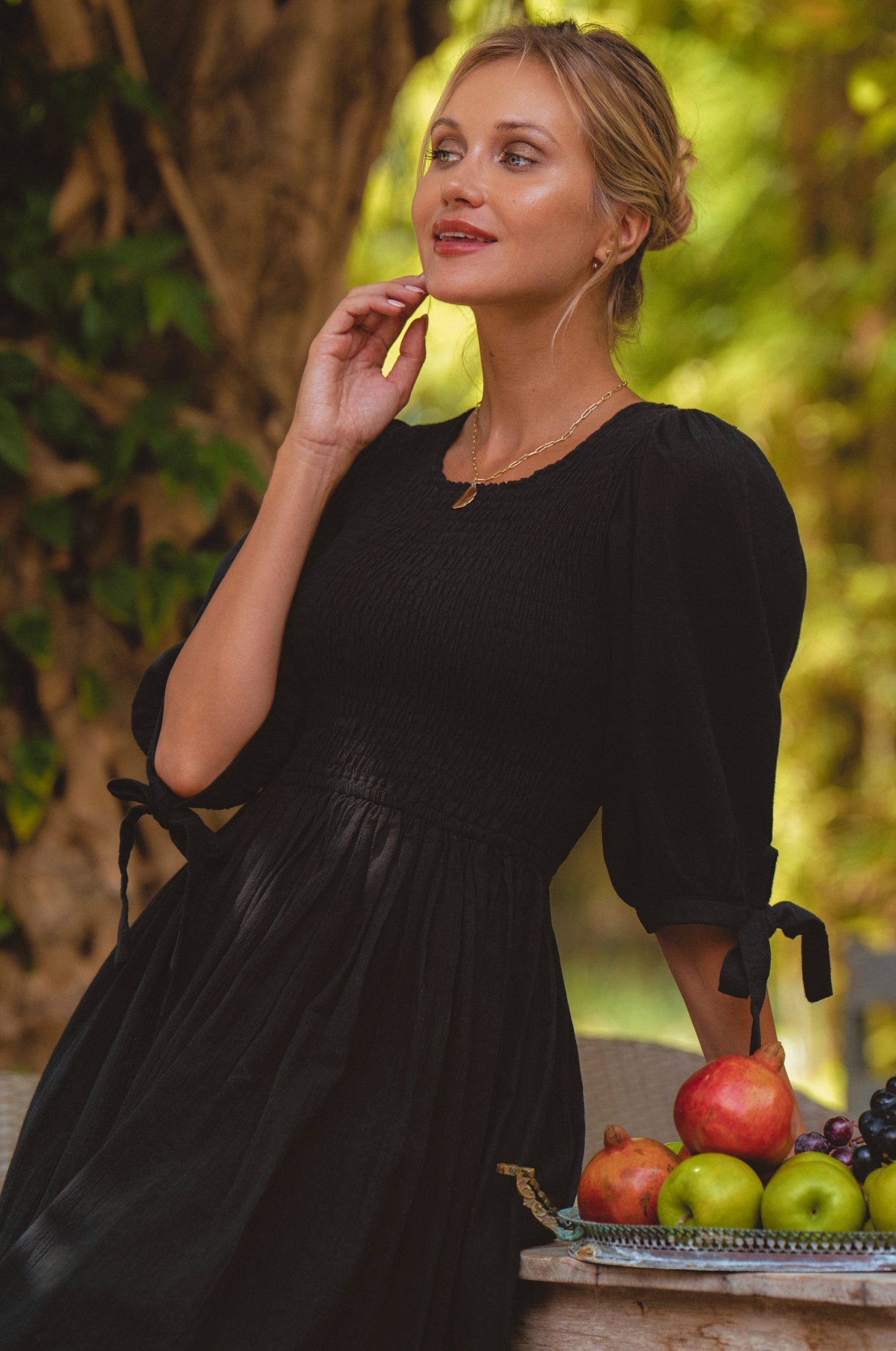 Woman in a black dress standing next to a table with fruits and vegetables outdoors.