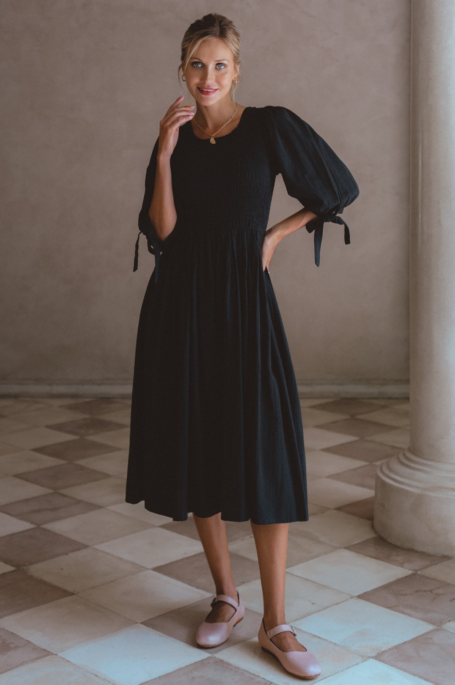 Woman wearing a black dress with smocked bodice, round neckline, and puffed sleeve standing in a room with a beige wall and tiled floor, front view