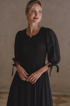 Woman wearing a black dress with smocked bodice, round neckline, and puffed sleeves against a neutral background
