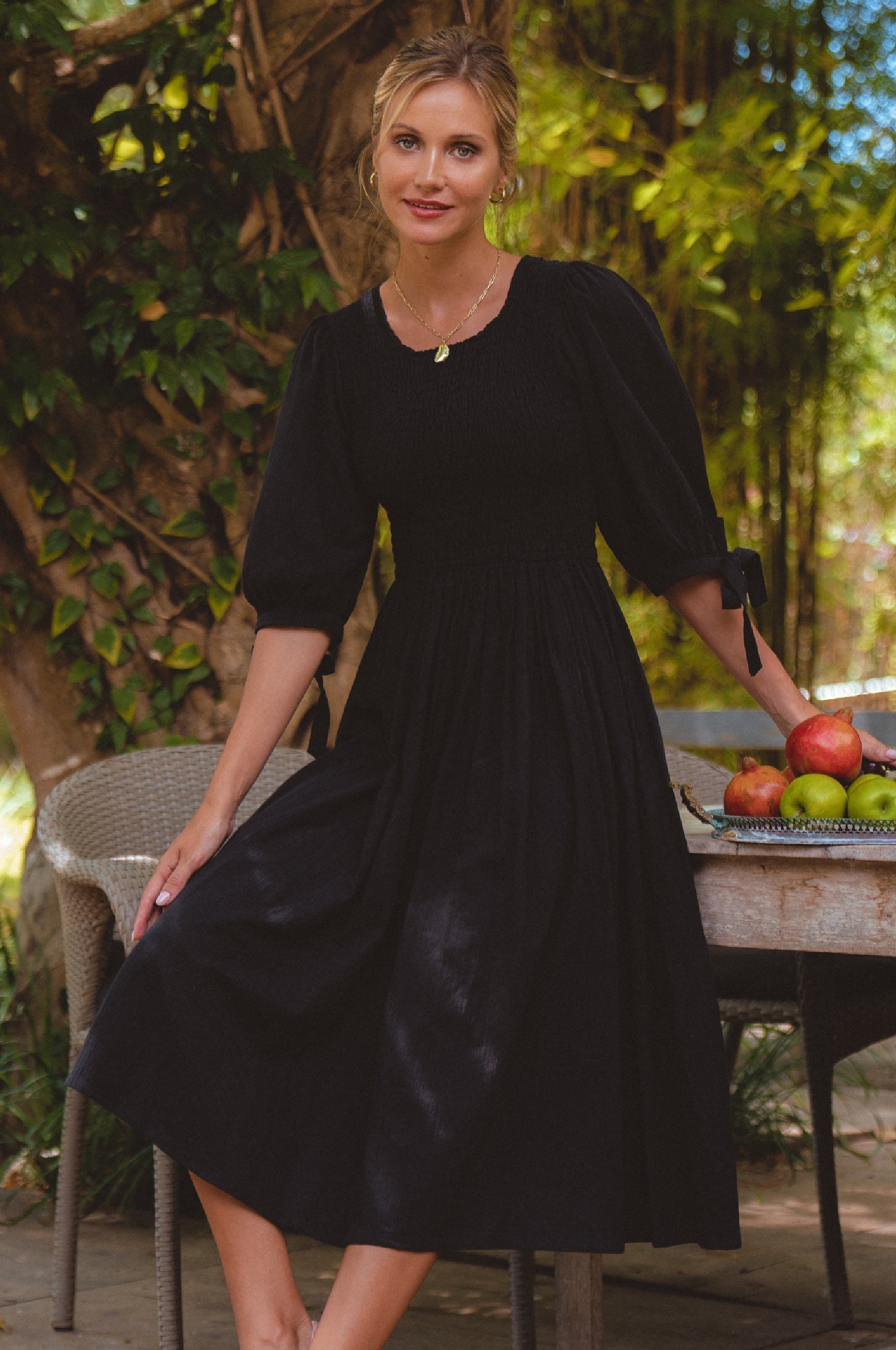 Woman in a black dress standing outdoors with greenery and apples on a table.