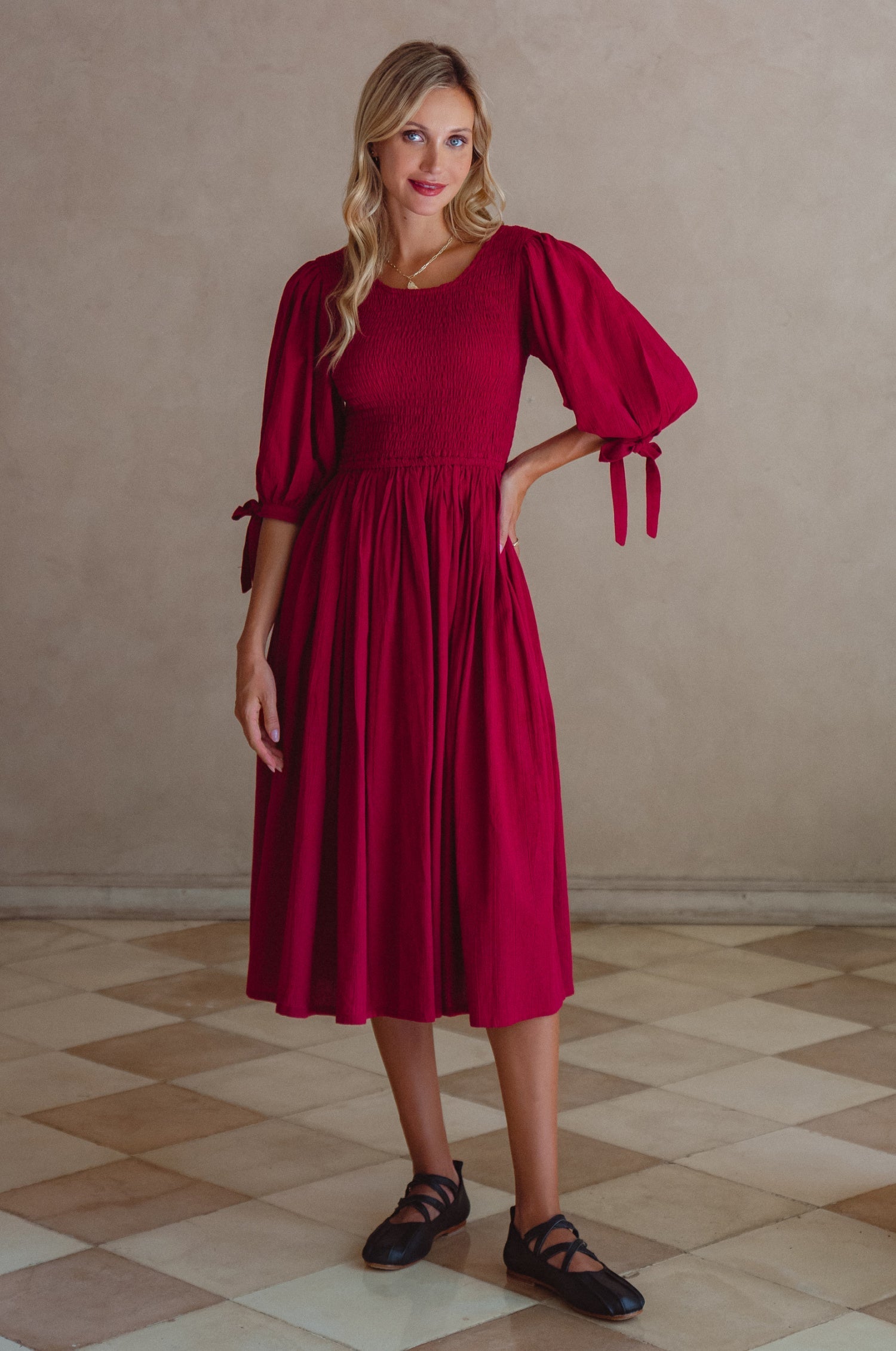 Woman wearing a red dress with smocked bodice, round neckline, and puffed sleeve standing against a beige wall, front view
