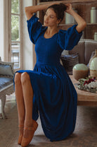 Woman in a blue dress with smocked bodice, round neckline, and puffed sleeve sitting on a couch in a living room.