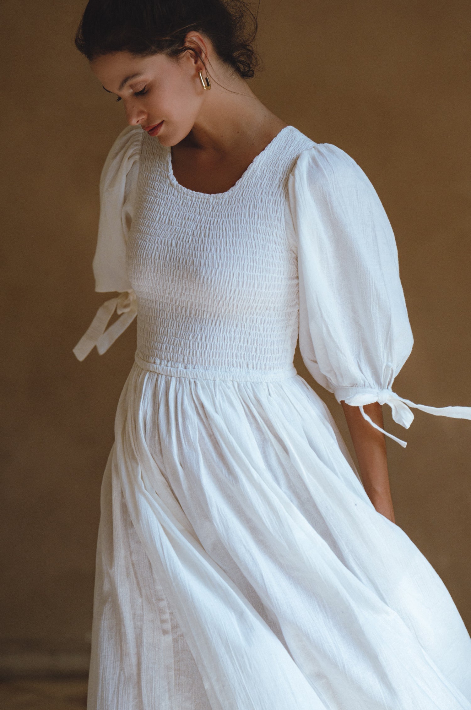 Woman wearing a white dress with smocked bodice, round neckline, and puffed sleeve against a brown background