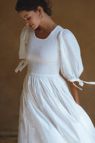 Woman wearing a white dress with smocked bodice, round neckline, and puffed sleeve against a brown background
