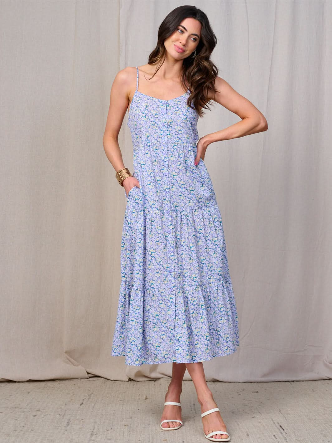 A woman wearing a floral maxi sundress with spaghetti straps, a scoop neckline, and tiered skirt. The dress is primarily soft purple with a floral print and she is posing against a neutral backdrop.