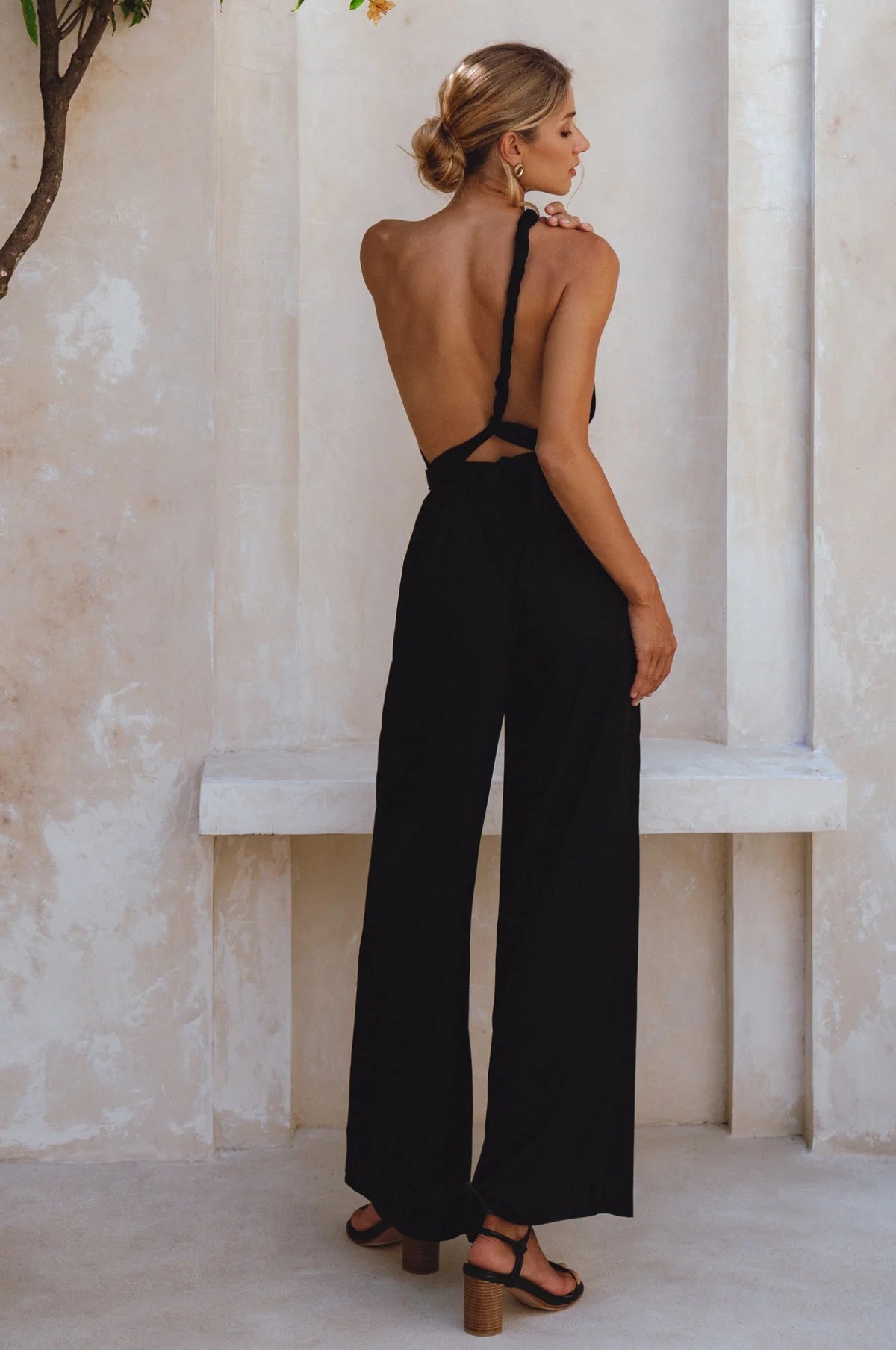 A woman stands with her back to the camera wearing a black open-back wide-leg jumpsuit with thin shoulder straps and cutout details, paired with brown block-heel sandals, set against a rustic beige stone wall and bench.