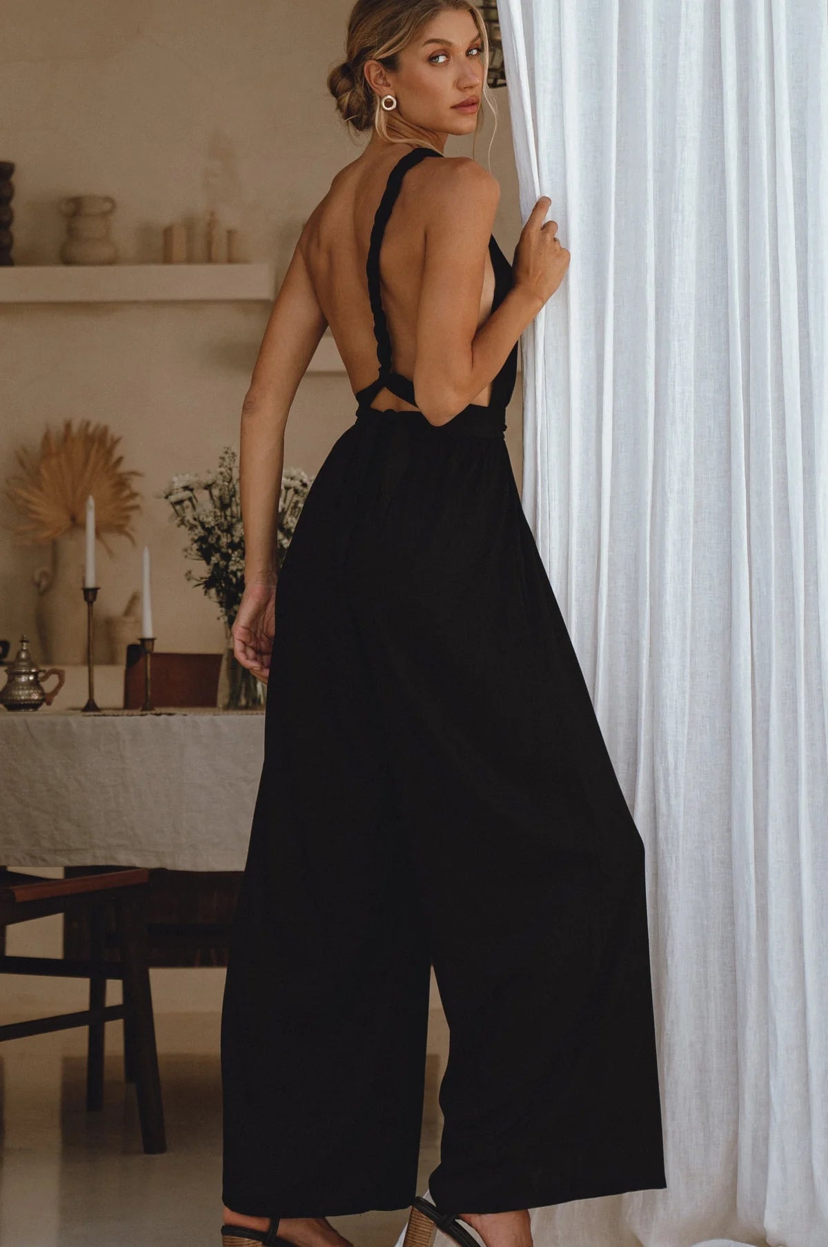 Woman wearing a black jumpsuit with a back cutout in a room with white curtains and a table