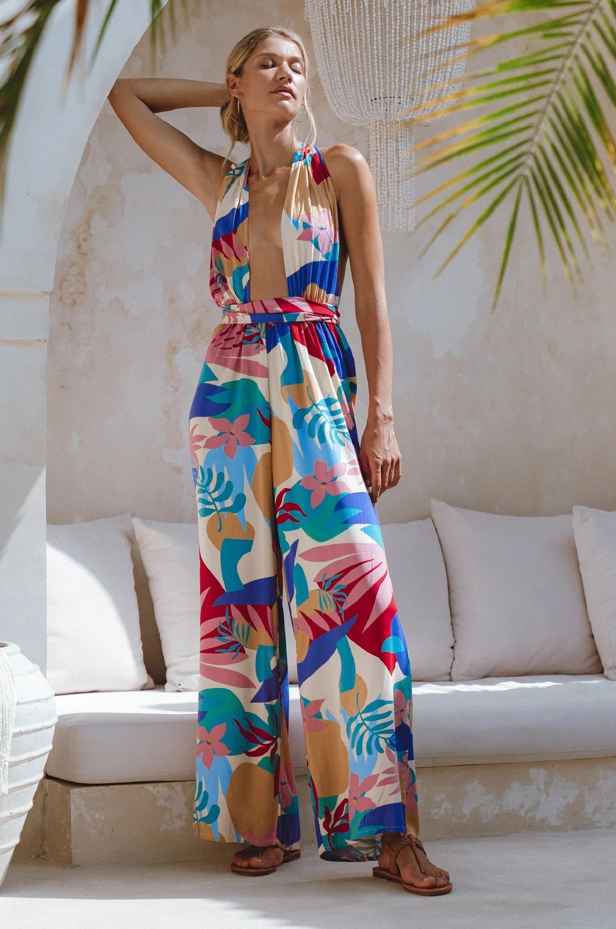 Woman wearing a colorful floral jumpsuit in an outdoor setting with a white couch and greenery.
