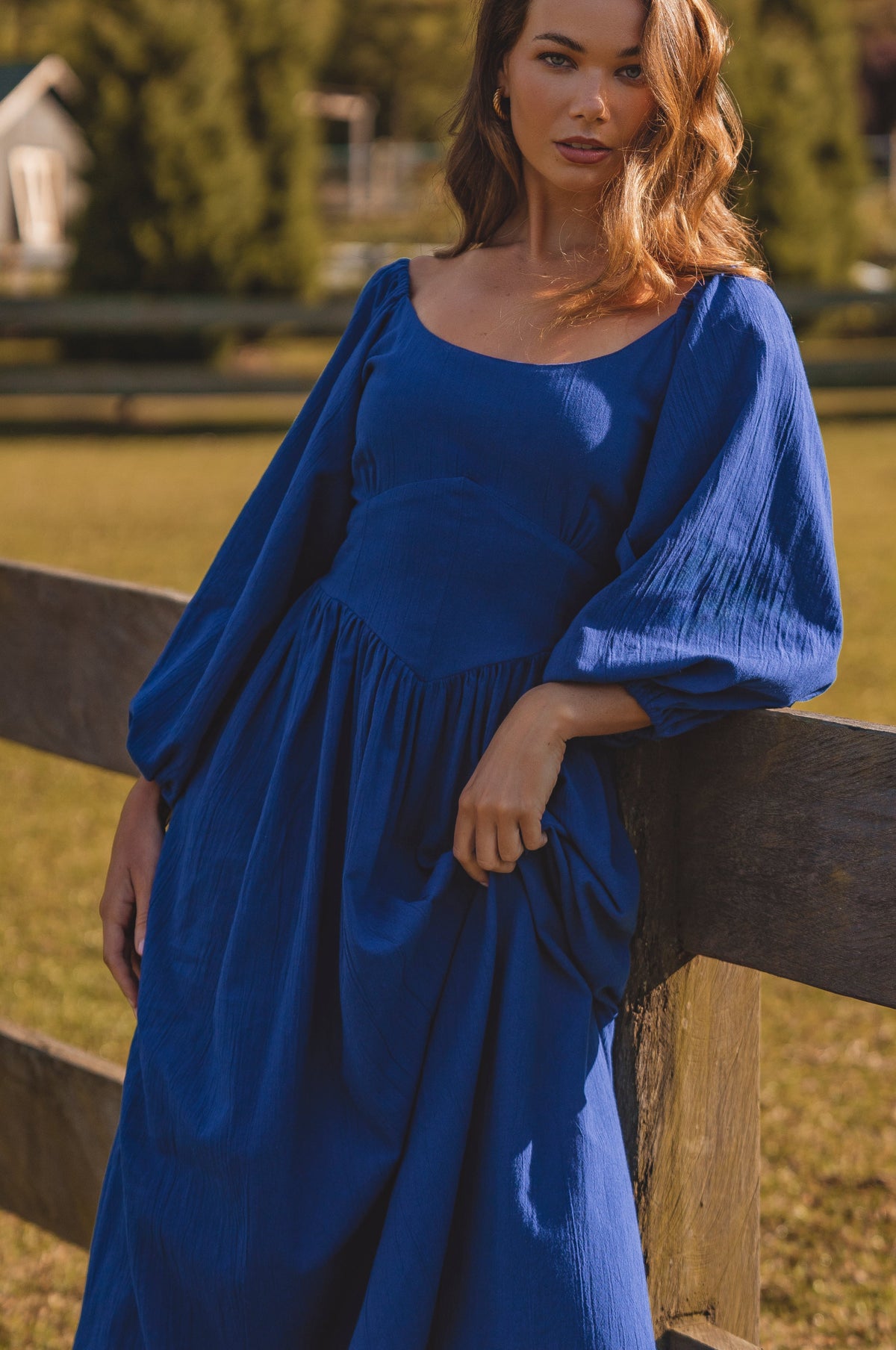 Woman wearing a blue dress standing by a wooden fence outdoors.