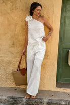 Woman in a white one-shoulder outfit holding a brown bag against a beige wall.
