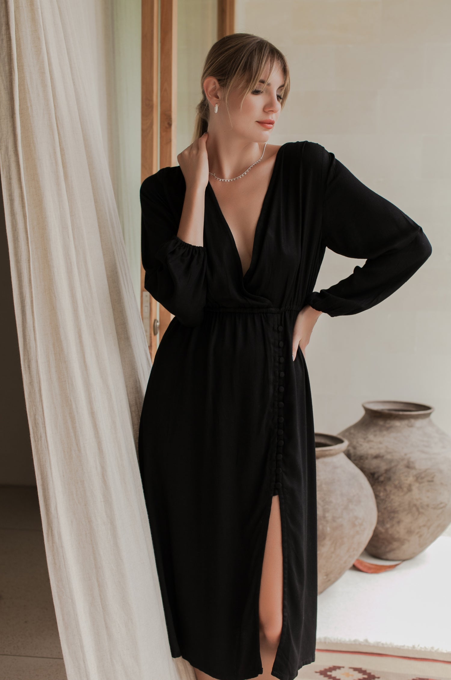 Woman wearing a black dress standing in a room with white curtains and decorative vases.