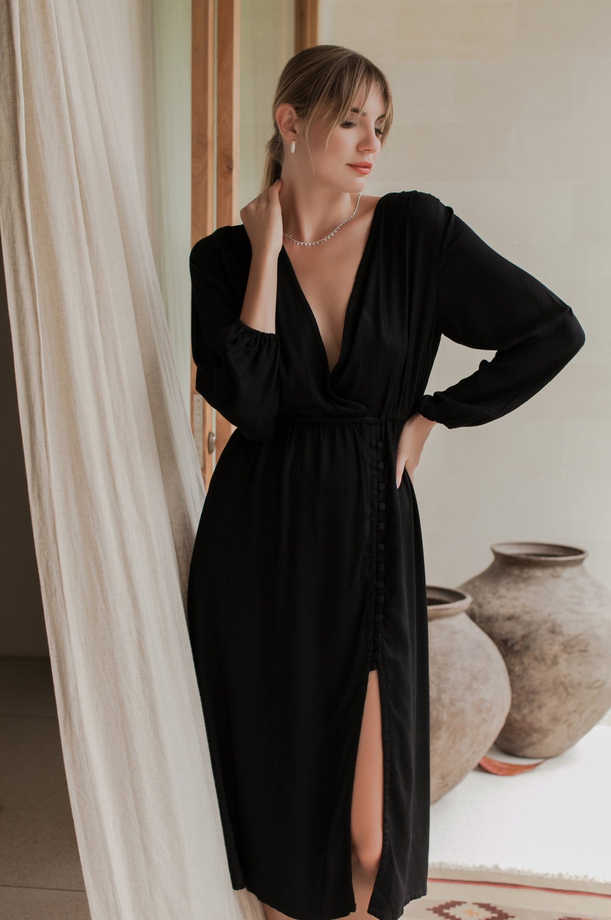 Woman wearing a black dress standing in a room with white curtains and decorative vases.
