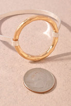 A gold-toned circular bracelet bangle with a white inner band, shown on a textured beige surface next to a US quarter coin for scale.