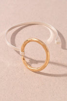 A minimalist cuff bracelet featuring a hammered gold circular ring connected to a smooth silver-toned open band, displayed on a textured beige background.