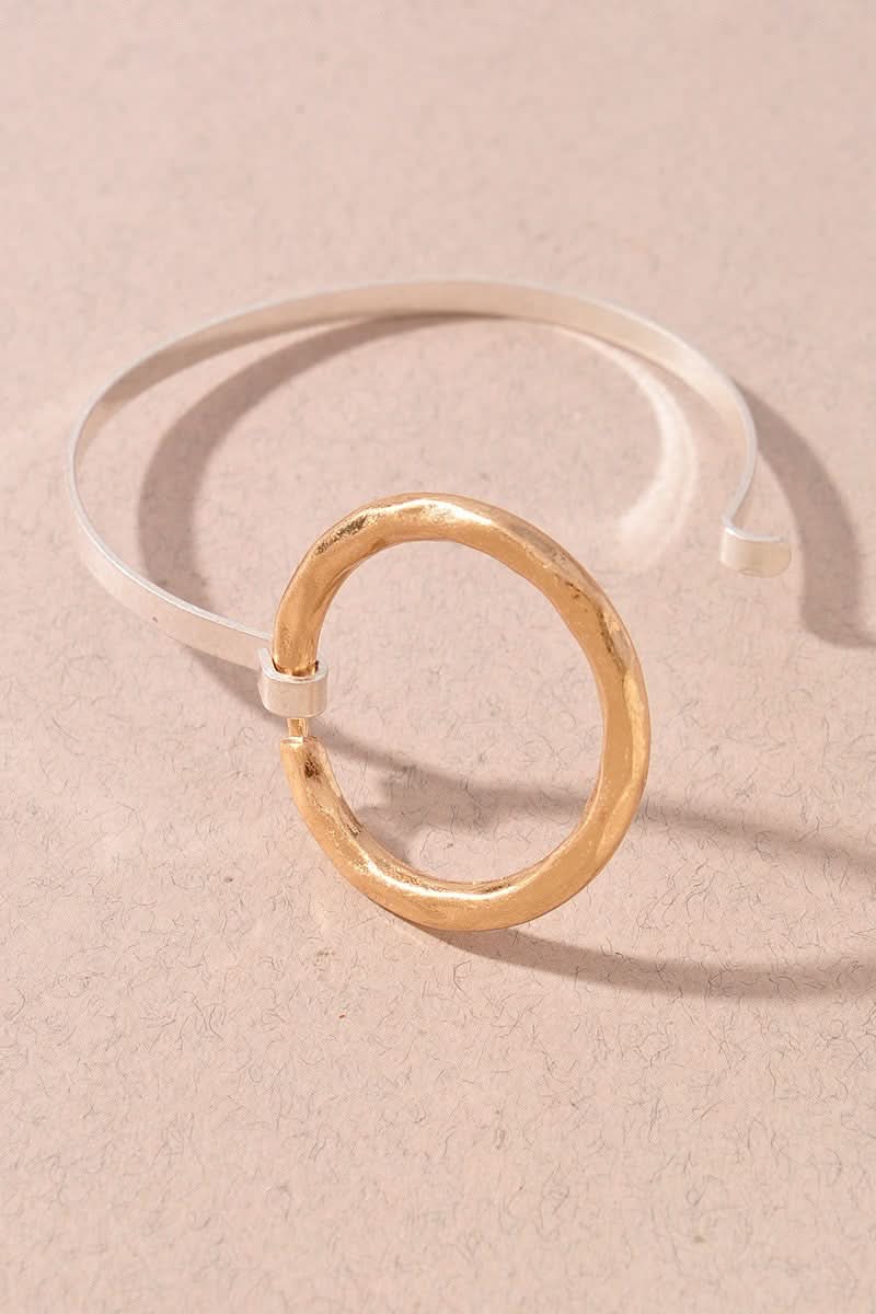 A minimalist cuff bracelet featuring a hammered gold circular ring connected to a smooth silver-toned open band, displayed on a textured beige background.