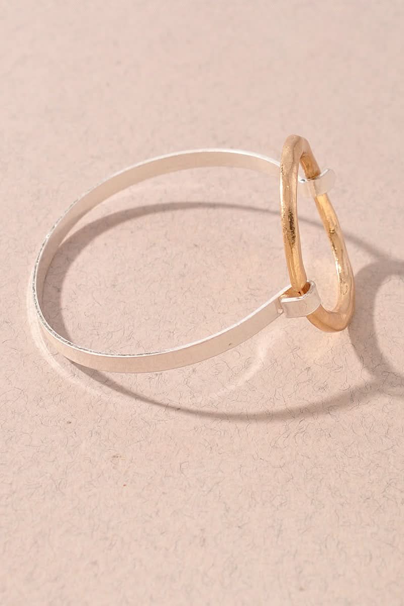 A minimalist two-tone bracelet featuring a thin silver band and a textured gold hoop, resting on a textured beige surface.