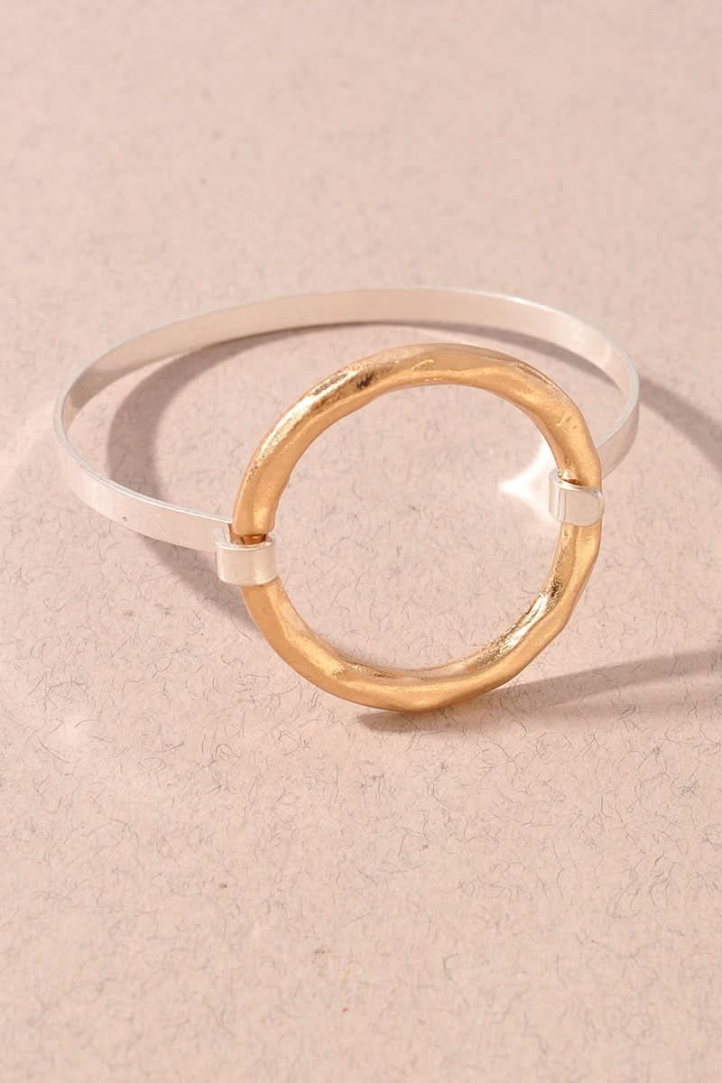 A minimalist bracelet bangle featuring a hammered gold circular disc overlaid on a polished silver band, displayed on a textured beige background.