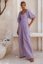 Woman wearing a lavender linen jumpsuit with puff sleeves and round neck standing against a neutral wall, 3/4 view