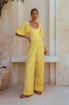 Woman wearing a yellow linen jumpsuit with puff sleeves and round neck standing against a neutral wall, front view