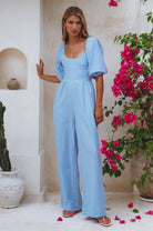 Woman wearing a lavender linen jumpsuit with puff sleeves and round neck standing against a neutral background with pink flowers, front view