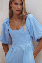 Woman wearing a light blue linen jumpsuit with puff sleeves and round neck standing against a plain wall, front view close up of bodice
