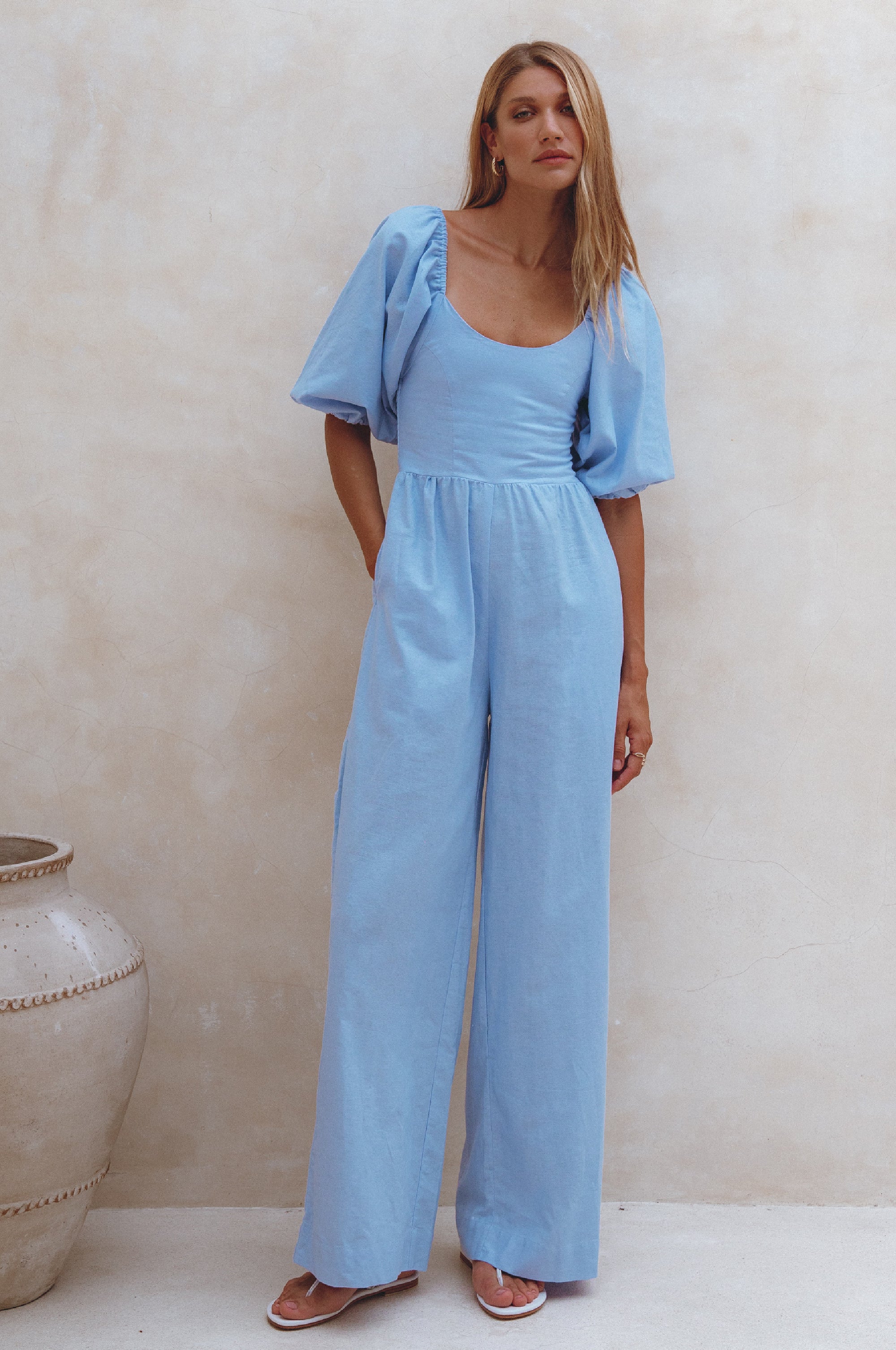 Woman wearing a light blue linen jumpsuit with puff sleeves and round neck standing against a plain wall.