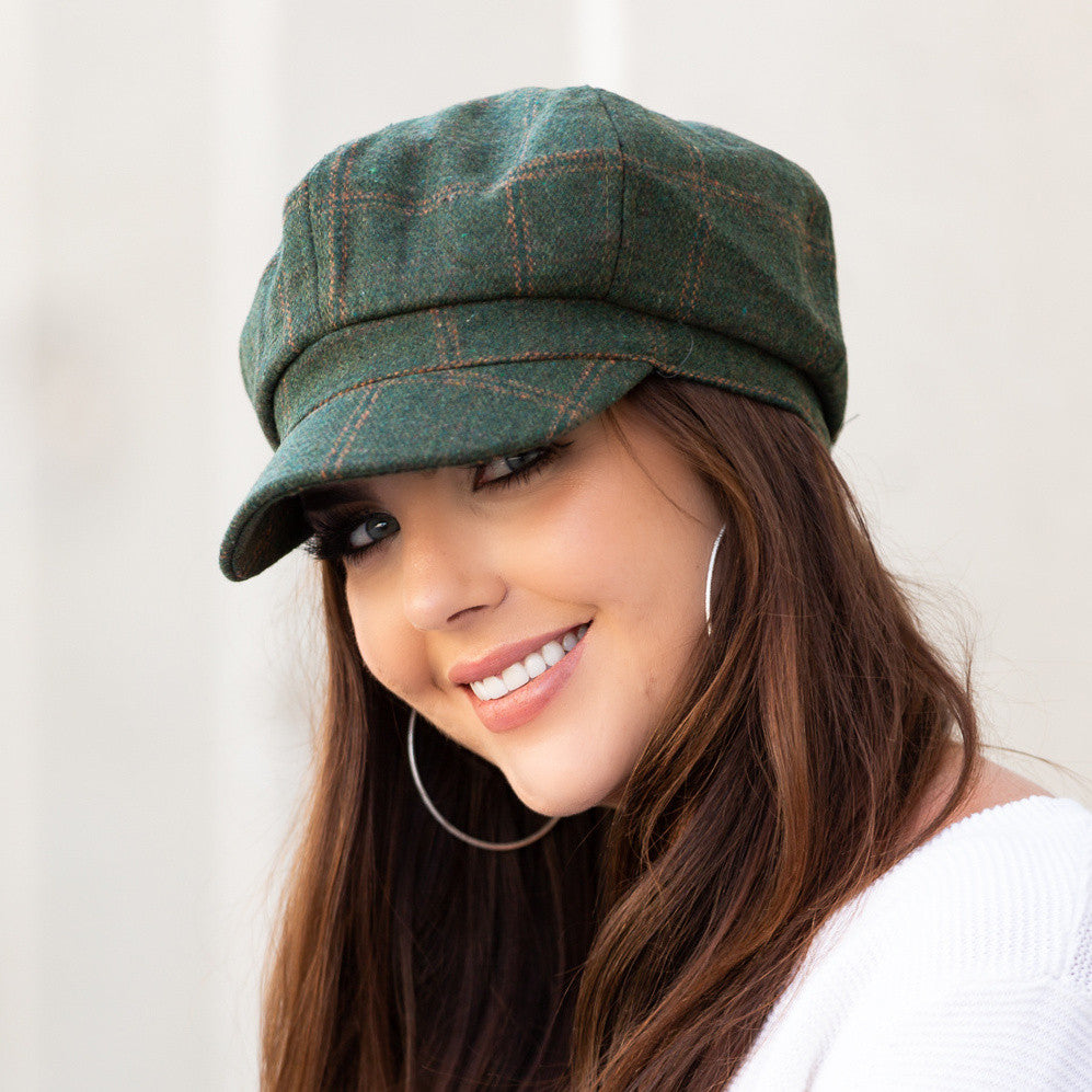 Woman wearing a hunter green tweed baker boy cap with a structured brim, side view against a pale wall
