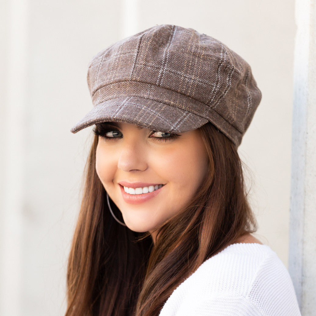 Woman wearing a tan tweed baker boy cap with a structured brim, side view against a pale wall