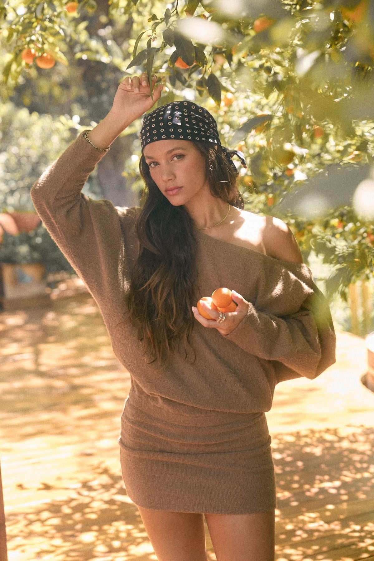 Woman wearing brown asymmetrical long-sleeve mini dress outdoors in a sunlit orchard.