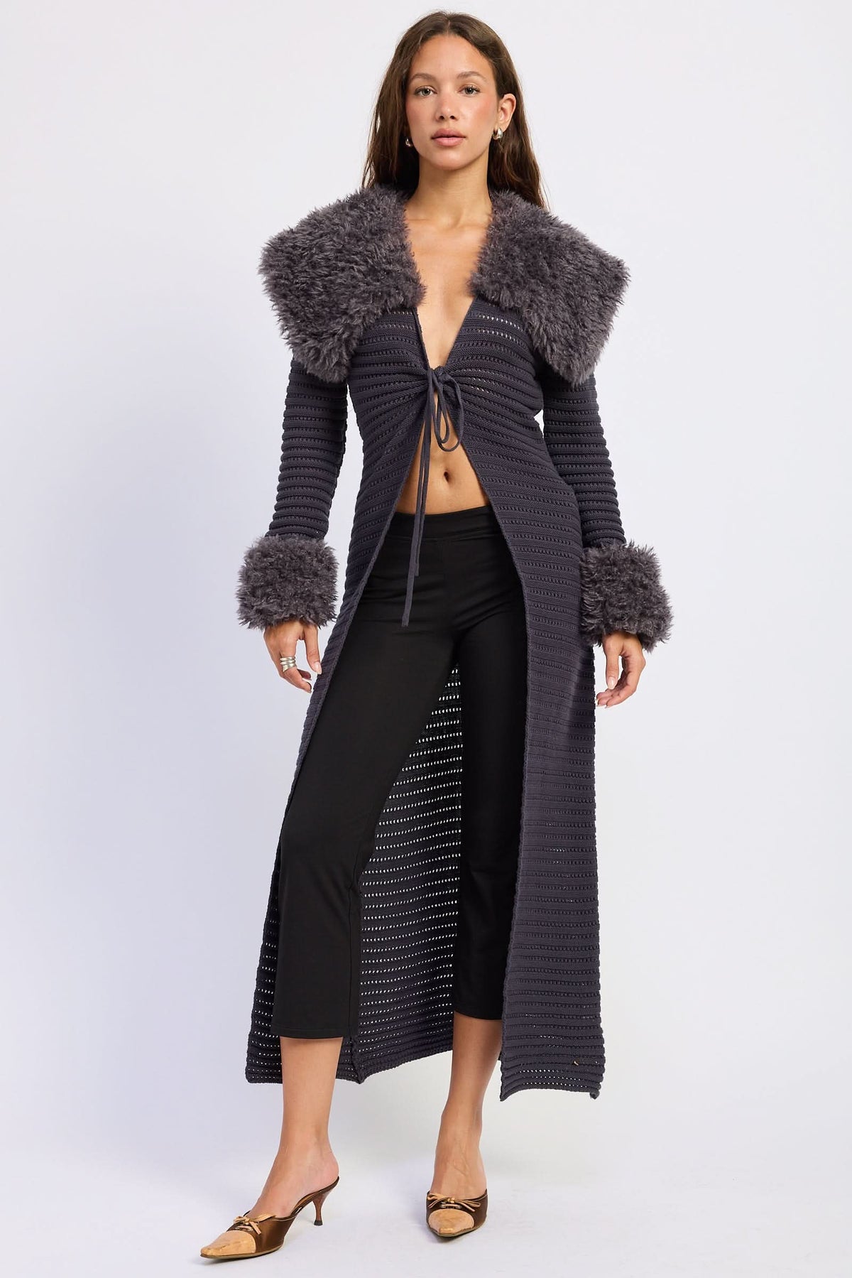 Woman wearing grey longline knit cardigan with plush collar, cuffs and front tie against white background.