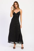 Woman wearing black V-neck maxi dress with delicate straps and floral detail against white background.