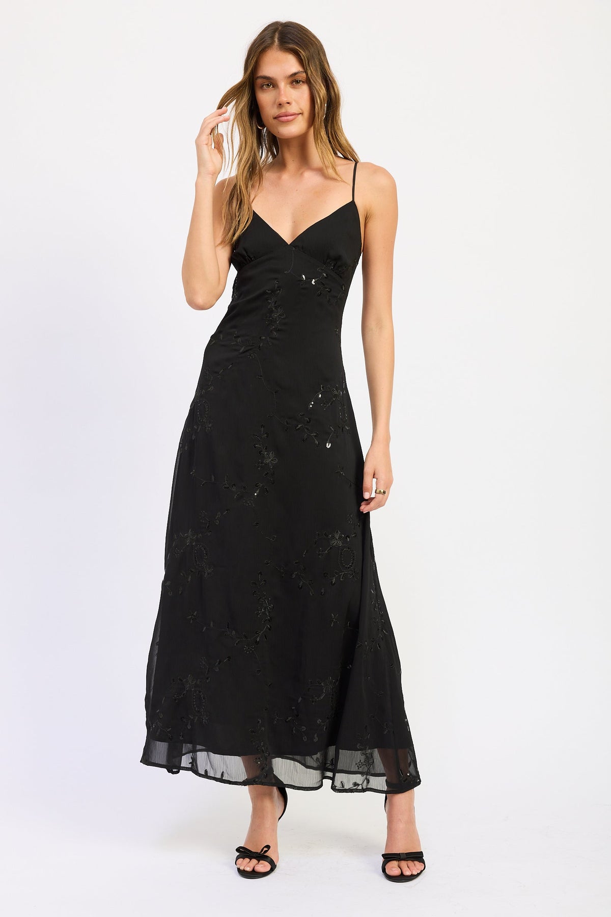 Woman wearing black V-neck maxi dress with delicate straps and floral detail against white background.
