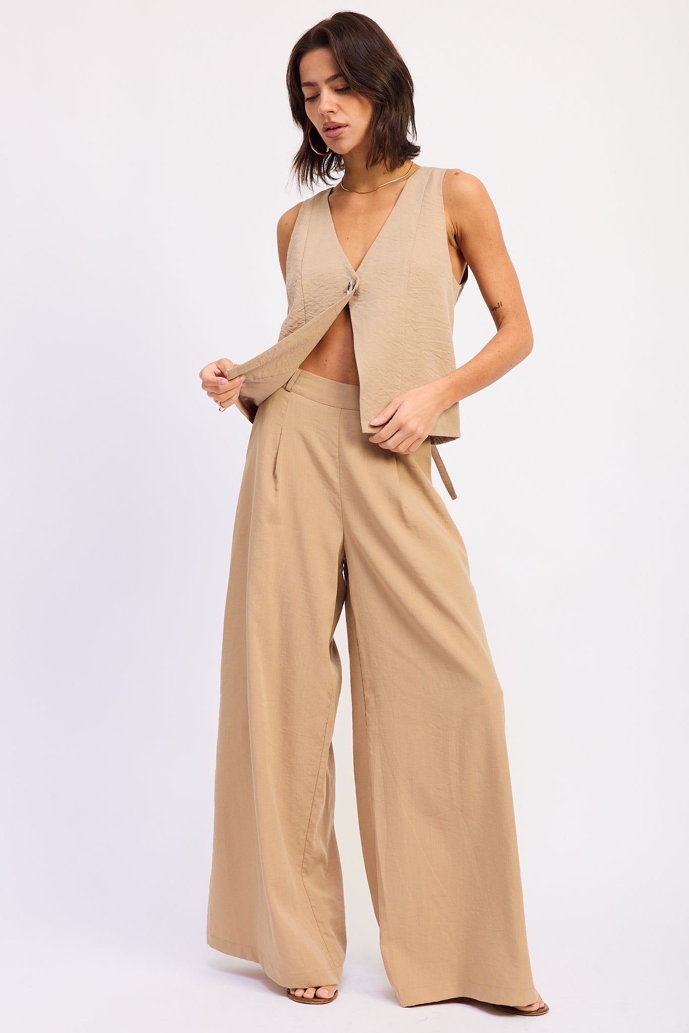 Woman wearing a Coffee sleeveless vest top with button front and tie back detail and wide-leg pants on a white background