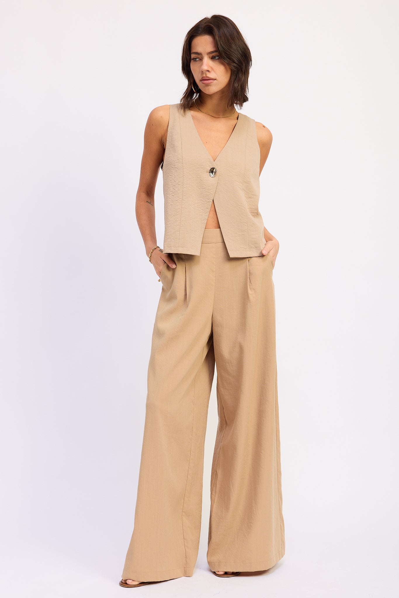 Woman wearing a Coffee sleeveless vest top with button front and tie back detail and wide-leg pants on a white background