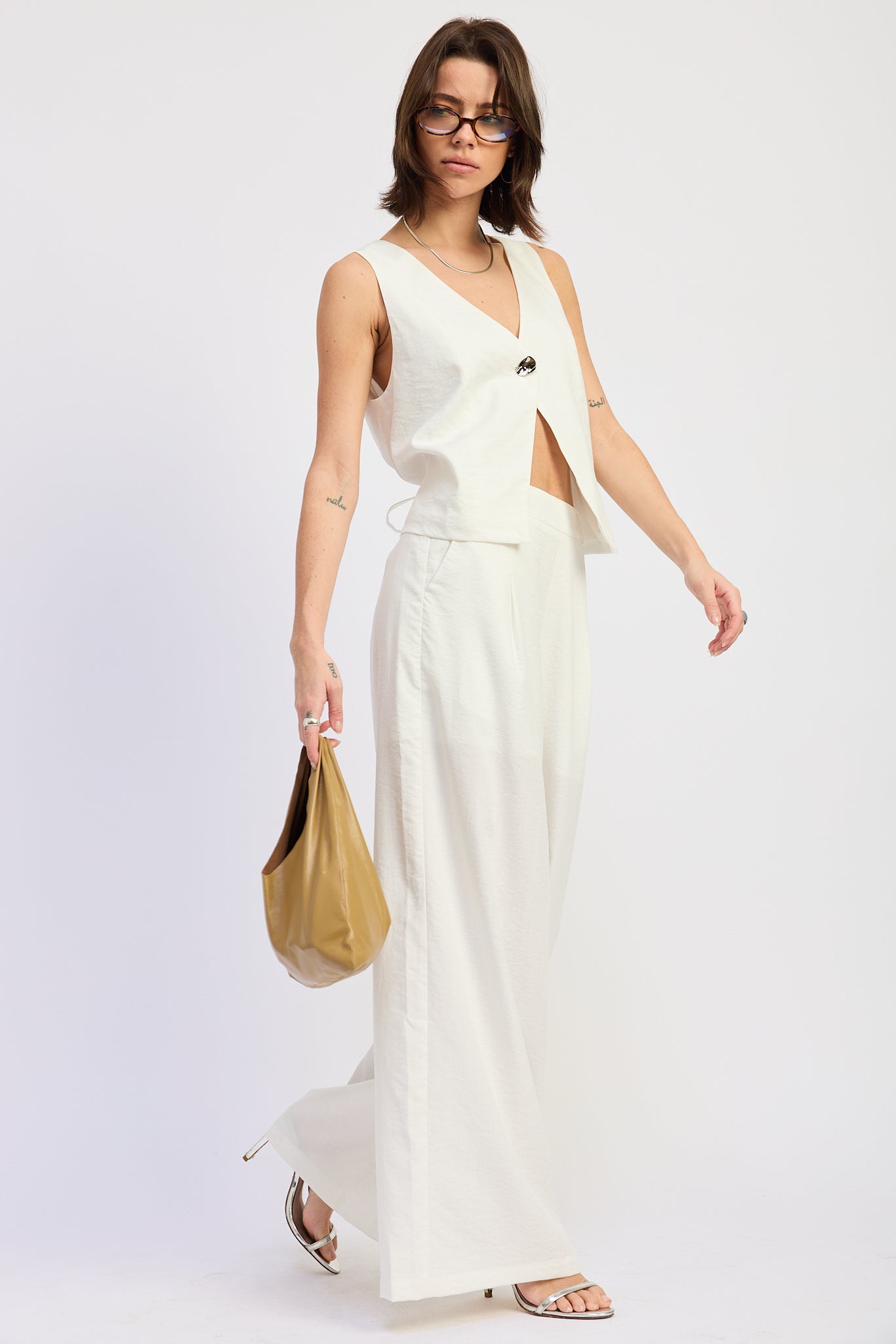 Woman wearing a white sleeveless vest top with button front and tie back detail and wide-leg pants on a white background