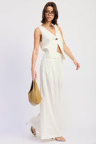 Woman wearing a white sleeveless vest top with button front and tie back detail and wide-leg pants on a white background