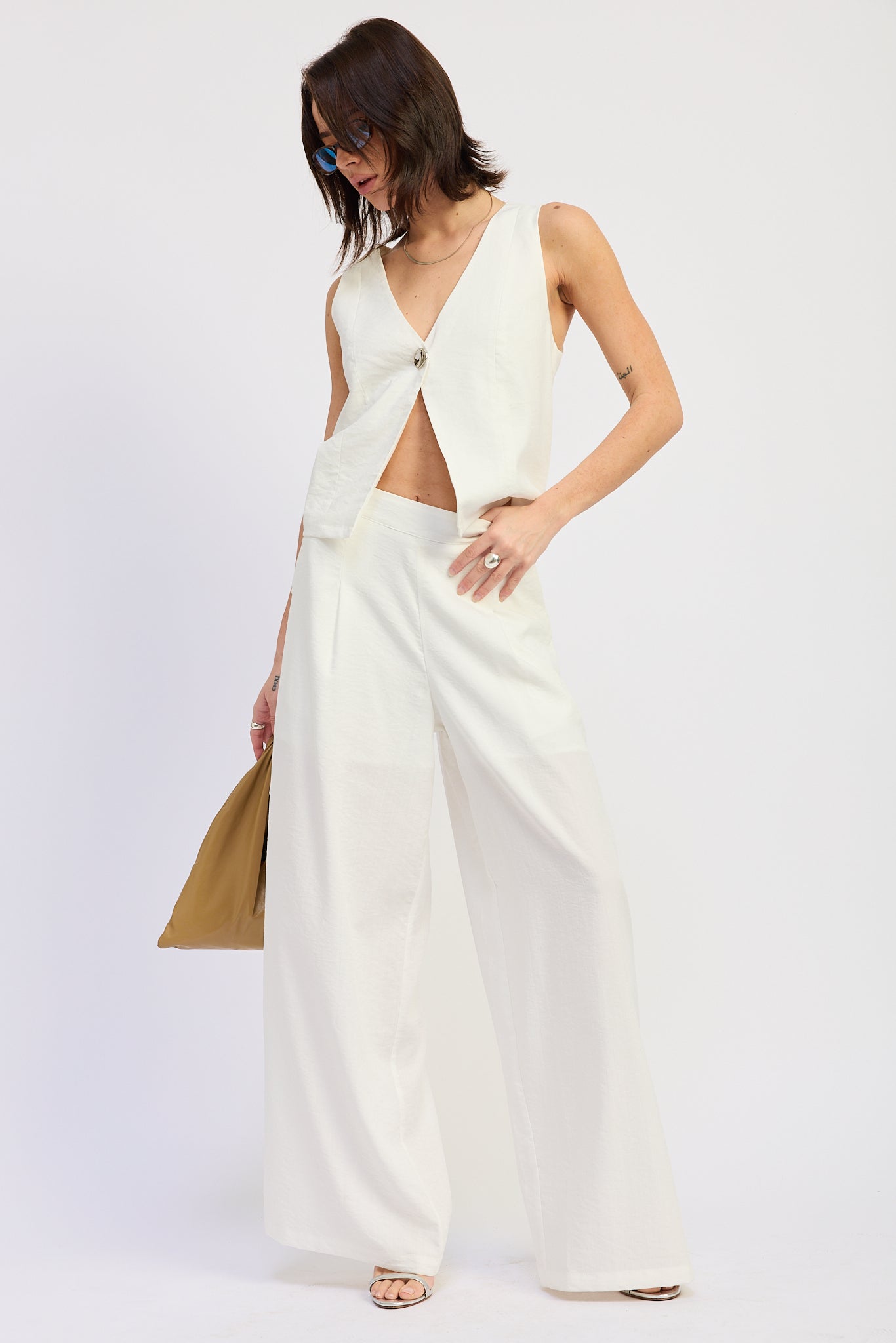 Woman wearing a white sleeveless vest top with button front and tie back detail and wide-leg pants on a white background