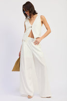 Woman wearing a white sleeveless vest top with button front and tie back detail and wide-leg pants on a white background