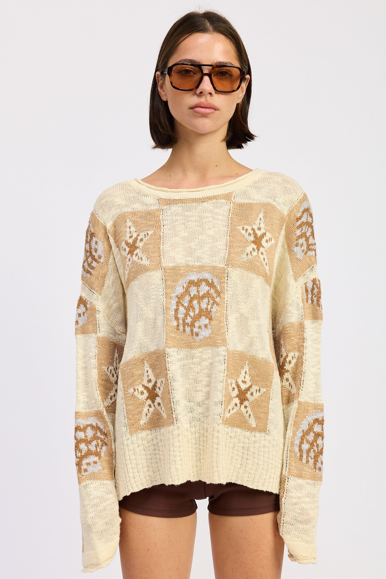 Woman wearing a cream and taupe patchwork knit sweater with star and seashell motifs, front view. and brown knee-high boots on a white background