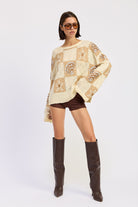 Woman wearing a cream and taupe patchwork knit sweater with star and seashell motifs, front view. and brown knee-high boots on a white background