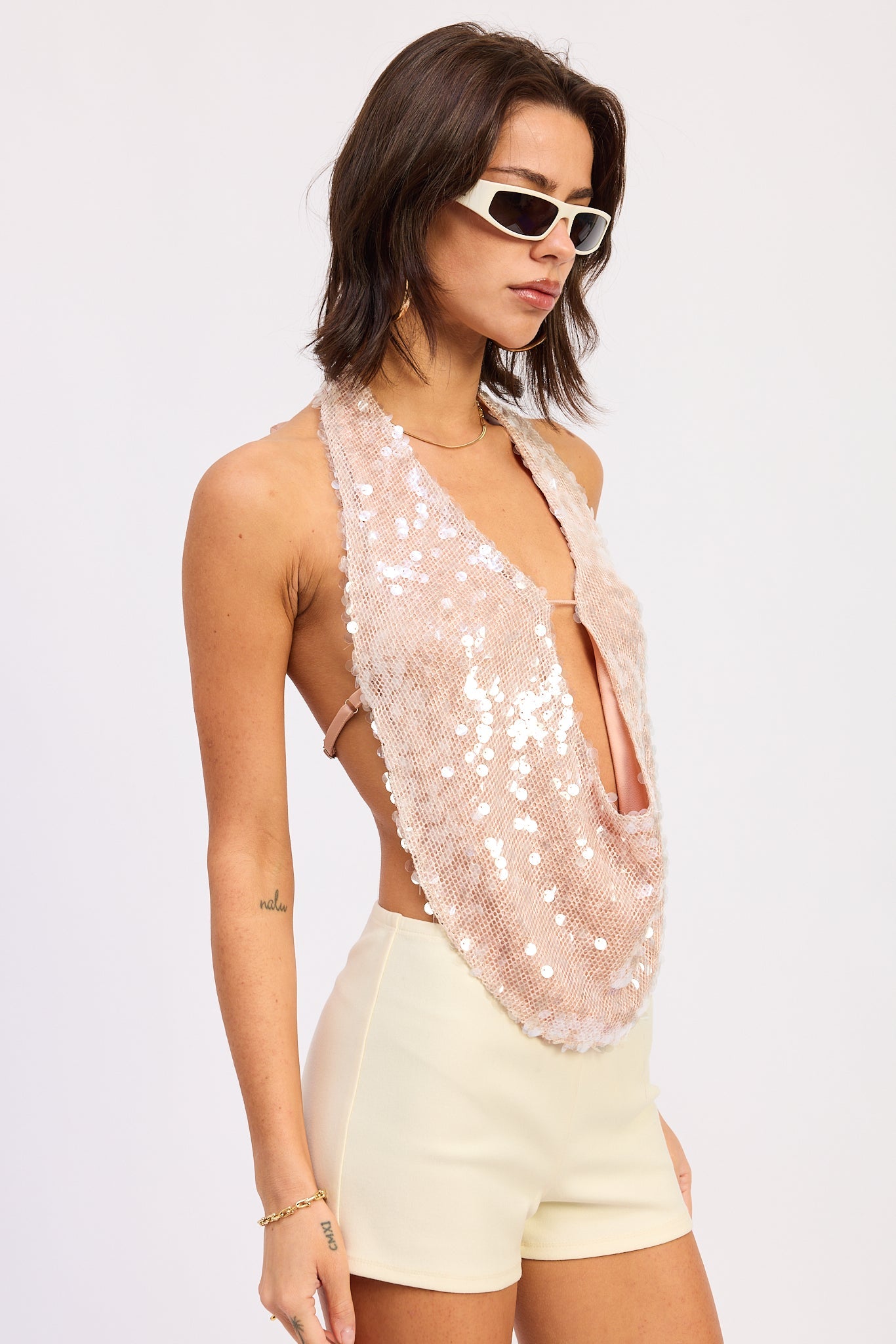 Woman wearing a pink sequin halter top and white shorts on a plain background side view