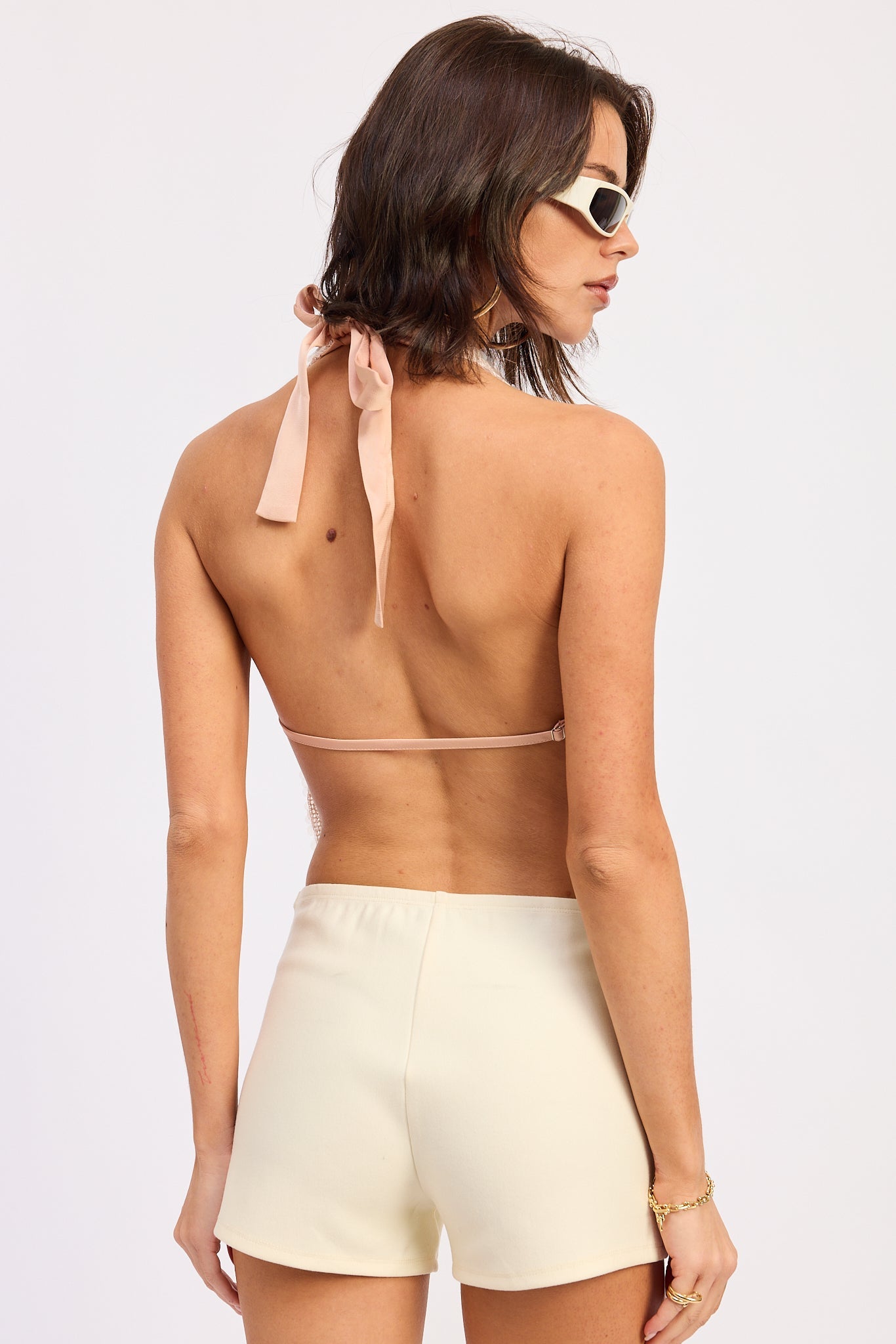 Woman wearing a pink sequin halter top and white shorts on a plain background back view