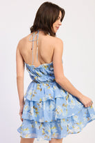 Woman wearing a blue floral ruffle tiered mini dress against a white background back view