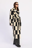 Woman wearing ivory and black checkered faux fur coat, side view showing belted waist against white background.