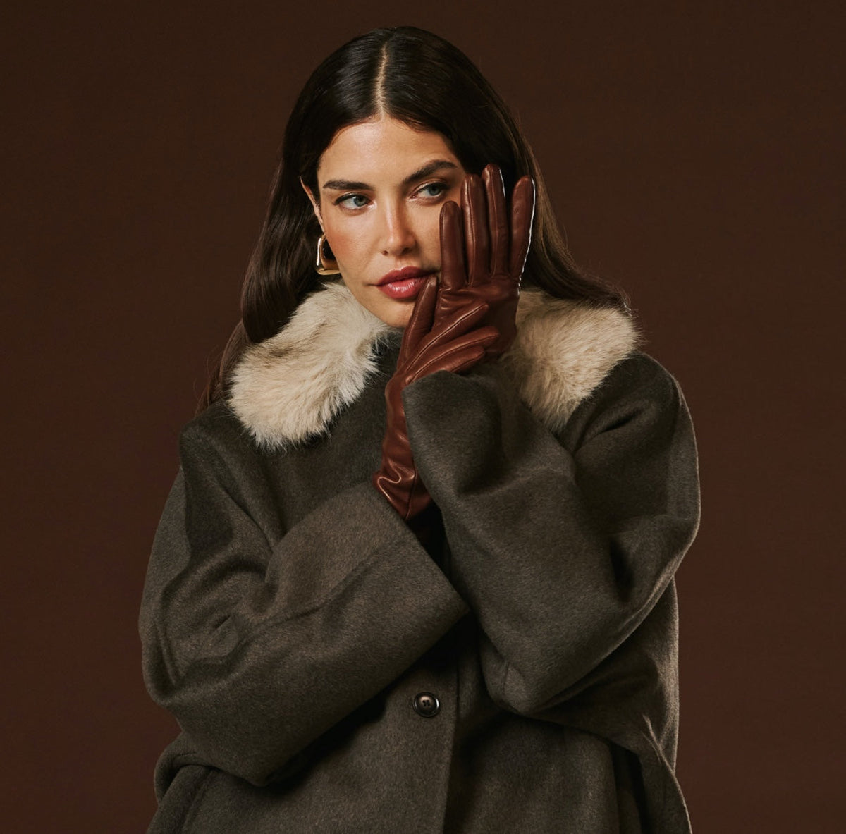 Woman wearing a dark coat with fur collar and brown leather gloves against a brown background