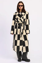 Woman wearing ivory and black checkered faux fur coat with wide collar and waist tie against white background.